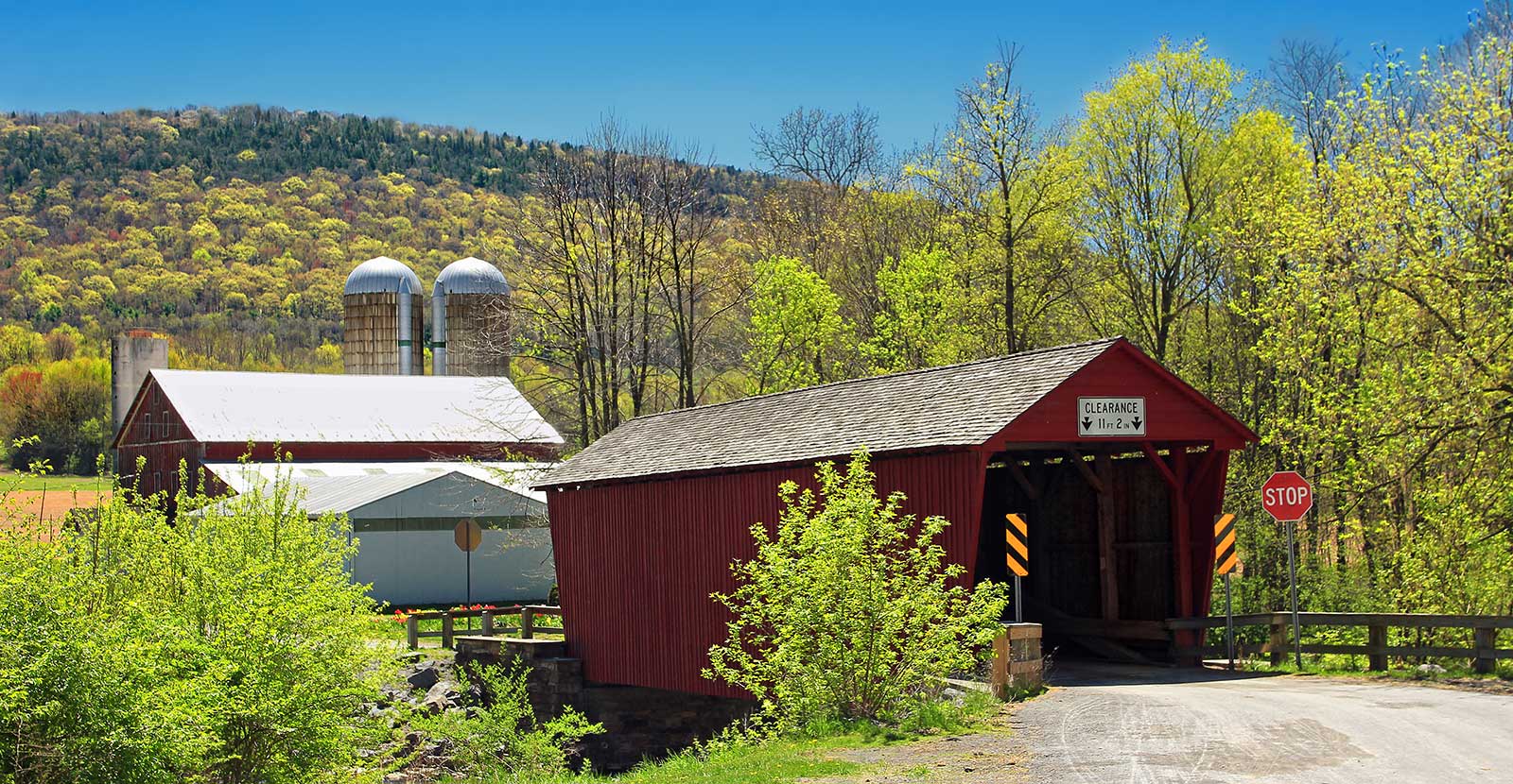 A covered bridge with a barn and silos in the background during spring. The leaves on the trees are new and bright green.